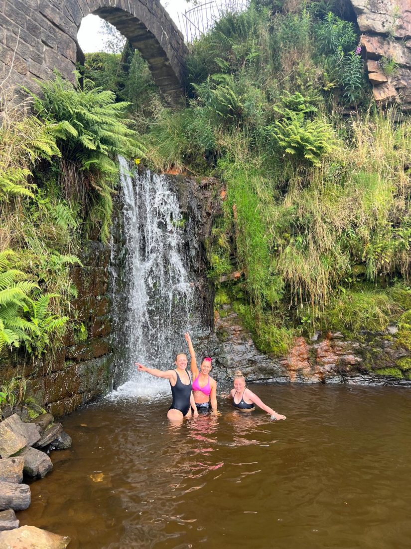 Three Solna swimmers in a plunge pool beneath a waterfall and an old stone arch