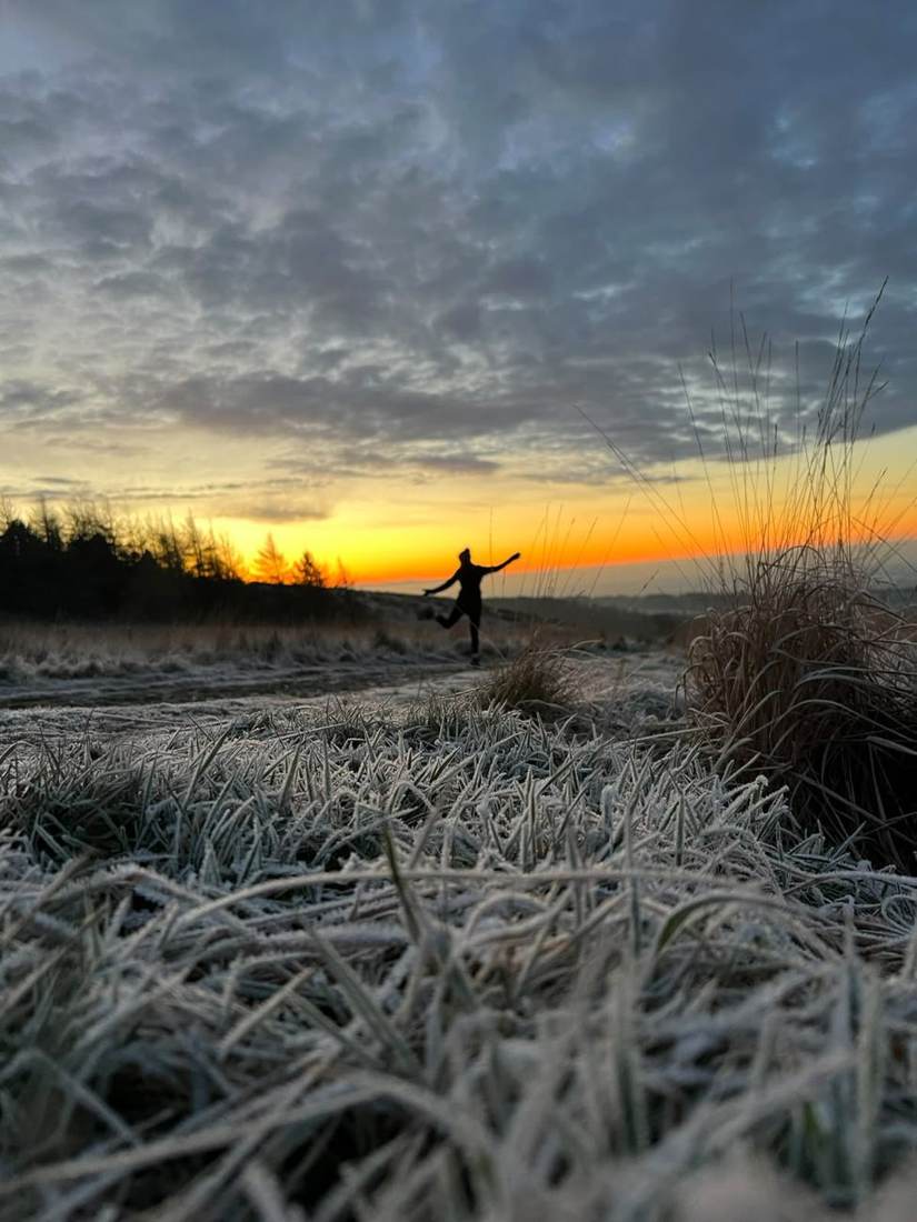 A walker jumping for joy against a winter sunset on frosted grass